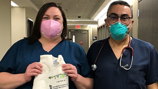 A woman and a man stand side by side with masks on. The woman is the nurse manager for the Birthing Center at UM Shore Medical Center at Easton and she is holding a HALO SleepSack, which is gifted to families at discharge.