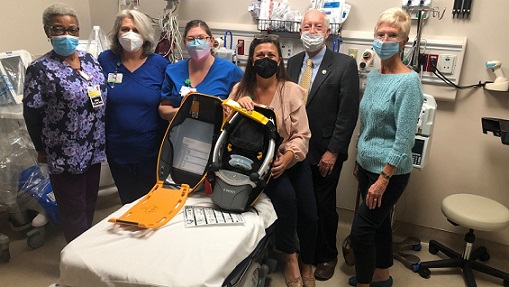 Five people stand around a woman who is sitting on a hospital bed holding a medical device that was just donated to UM Shore Medical Center at Cambridge.