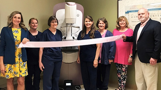 Seven people stand on either side of a brand new mammography machine. The one furthest on either side is holding a pink ribbon, which is draped in front of the other five people and the mammography machine.
