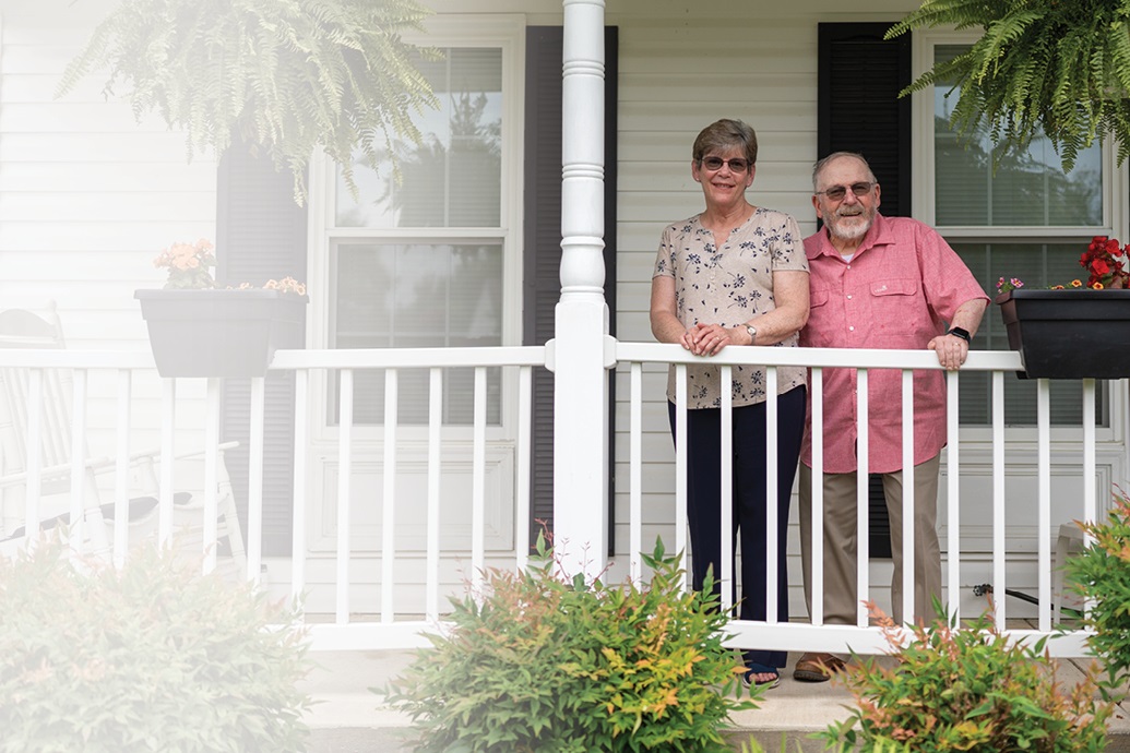 Cathy and her husband are standing on the porch smiling.